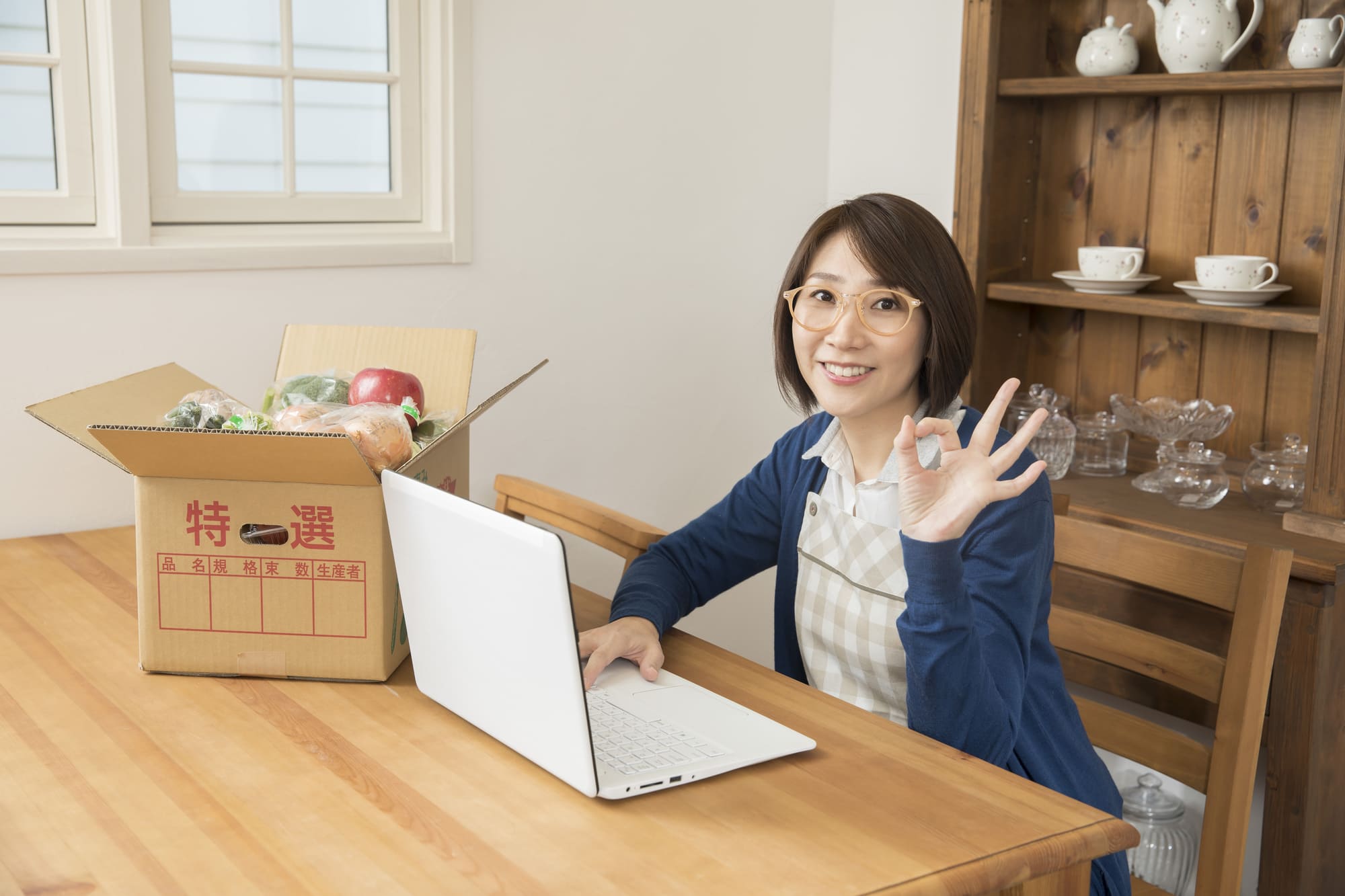 食材の入った段ボール箱とパソコンの前にいる笑顔の女性
